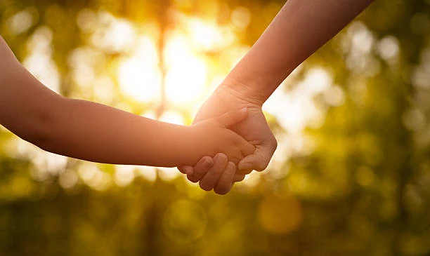 La main d’un adulte tenant la main d’un enfant, à contre-jour devant un feuillage.